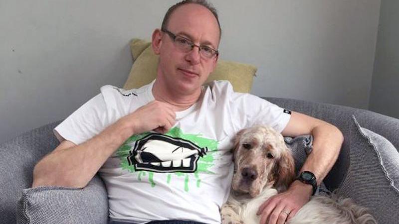 A man in glasses and a white t-shirt sits holding a dog
