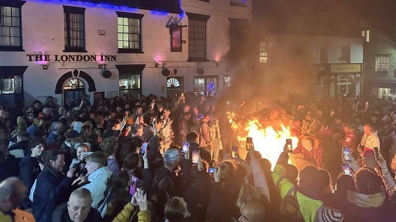 A large crowd on a street with their mobile phones out filming and photographing a flaming tar barrel being carried on someone's back.
