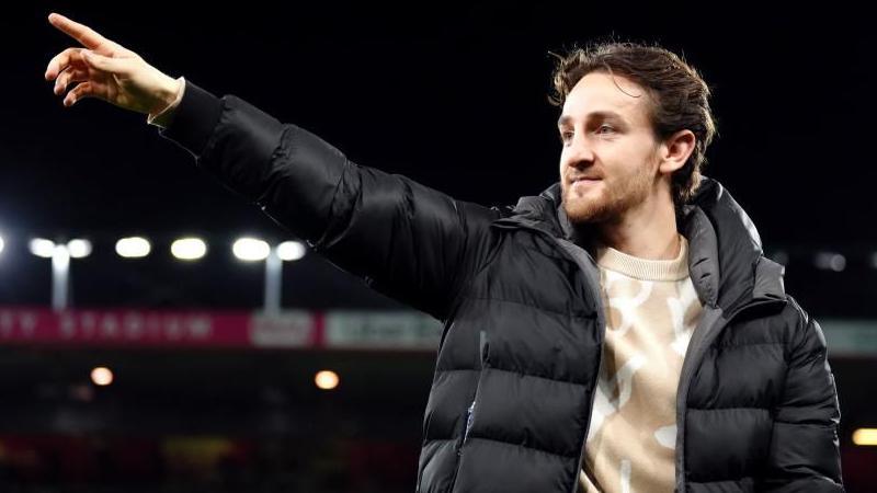 Tom Lockyer, a Luton football player, standing on a football pitch, wearing a black coat and beige jumper, pointing his finger to the crowd. He is smiling and has dark brown hair and a beard.