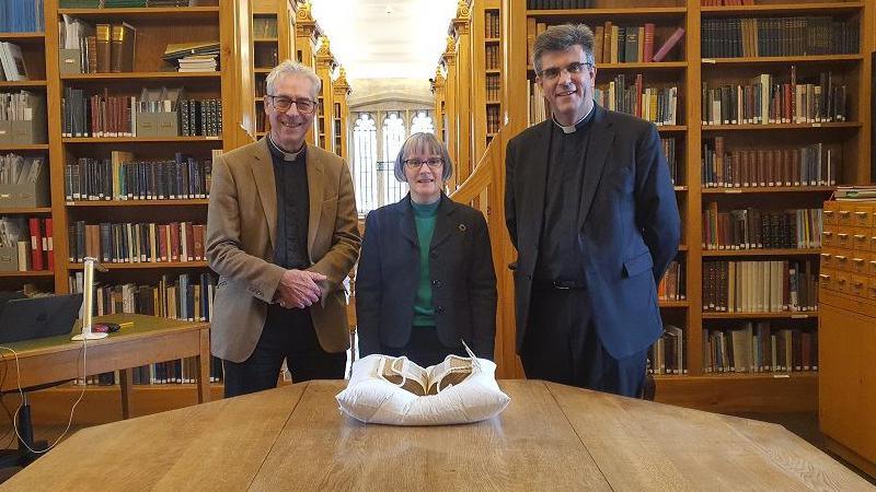 Canon Treasurer Ed Probert, librarian Dr Anne Dutton and The Revd Nicholas Papadopulos, Dean of Salisbury standing behind a table which has the manuscript on it. They are all looking at the camera and smiling, standing in the library, with rows of shelves behind them.