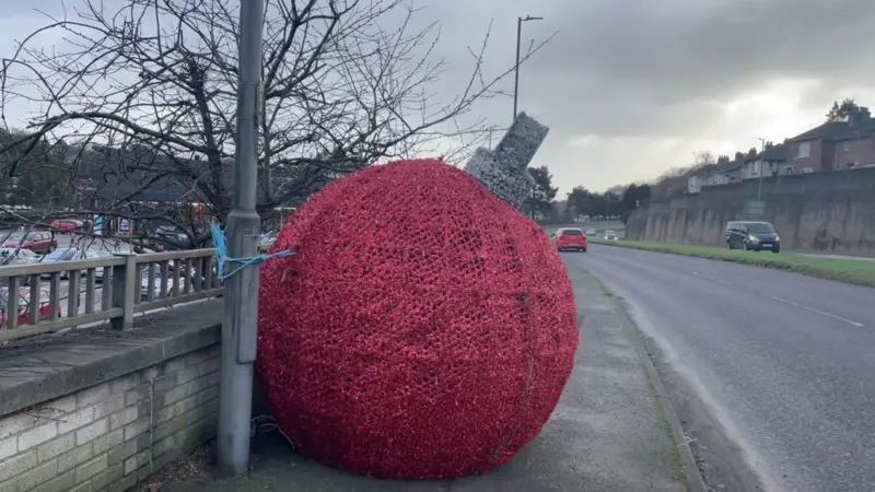 A giant red bauble Christmas decoration about 5ft in diameter sits on a pavement by a dual carriageway, fixed to a lamp post by a short blue cord.