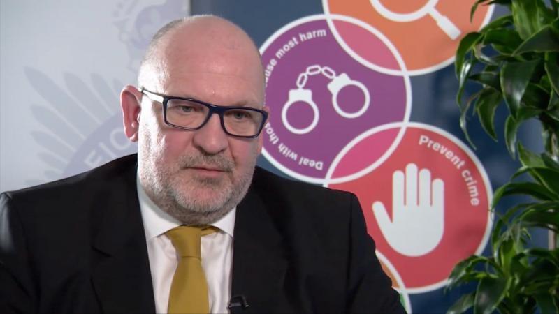 Inspektori i Përkohshëm i Detektimit, Neil Holden, me syze dhe kostum të zi me kravatë të verdhë.