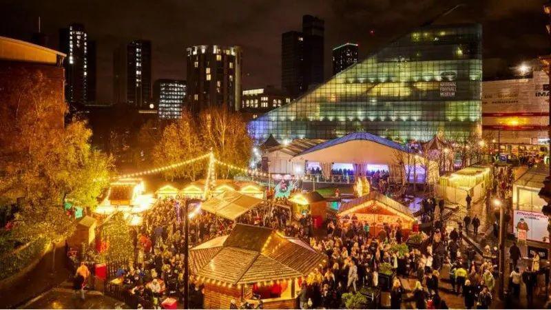 The stalls of Manchester's Christmas market seen from above, lit up in with yellow lights from above at night, with skyscrapers seen in the background and crowds of people filling the street. 