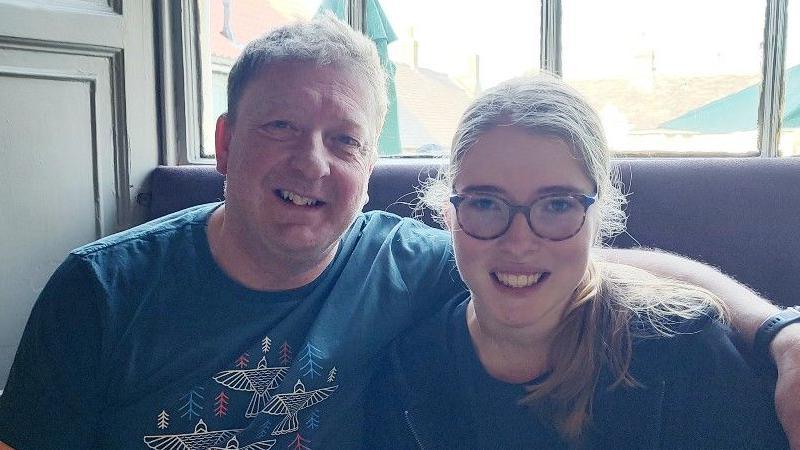 Jess Lowe is sitting in the right of the picture. She is with her dad in what appears to be a restaurant. Her dad has his arm around her shoulder and they are both smiling. Outside the window behind them, a number of green parasols can be seen. 