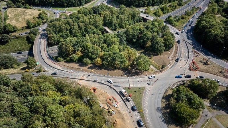 The Queen Eleanor roundabout, showing a large roundabout, with lots of trees in the middle, trees all around it, work being carried out around it, cars on the roads and several junctions. It is an aerial shot.
