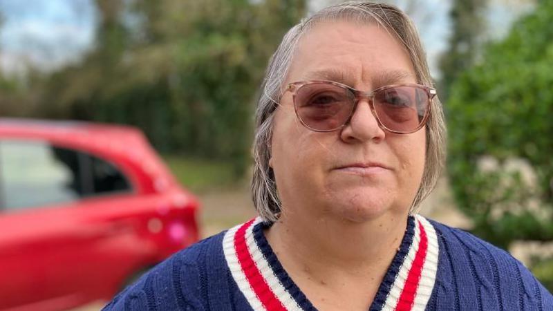 Debbie Barrett, looking straight at the camera, wearing a blue jumper with a V-neck and white, red and blue trim. She is wearing glasses and has chin length hair. A red car is behind her and a bush to the right, both blurred. 