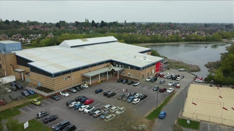 An aerial view of a big beige brick building with a grey roof, situated next to the waterfront. Lots of cars are parked in the car park. There are many red-brick houses on the horizon. 