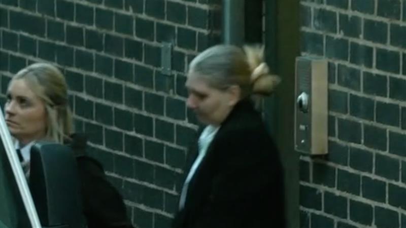 A blurry image of a woman with blonde hair tied up in a bun wearing a black coat leaving through the side door of a court, with dark grey bricks and a silver intercom. There is also a prison officer with blonde hair next to her