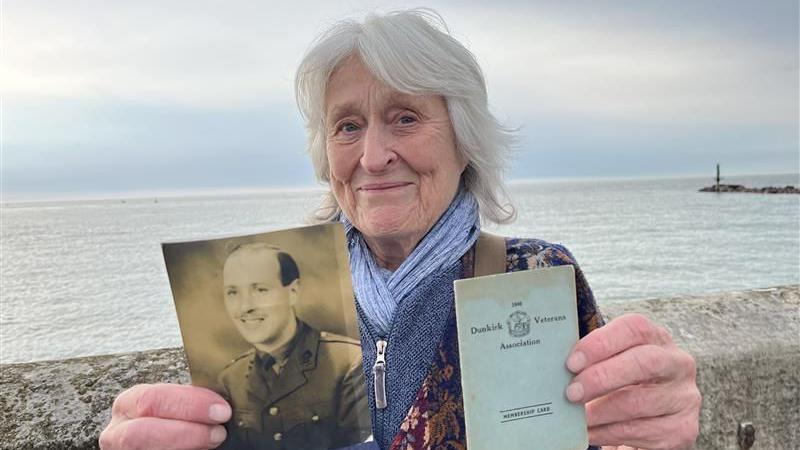 A woman with white hair smiling at the camera and standing by a wall with the sea behind her. She is holding a black and white picture of a man and a small book which says "Dunkirk Veterans Association" on the front.