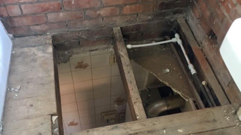 A bathroom floor with a hole in it, with bare wooden boards, exposed pipes and a view down to the tiles of the bathroom below. 