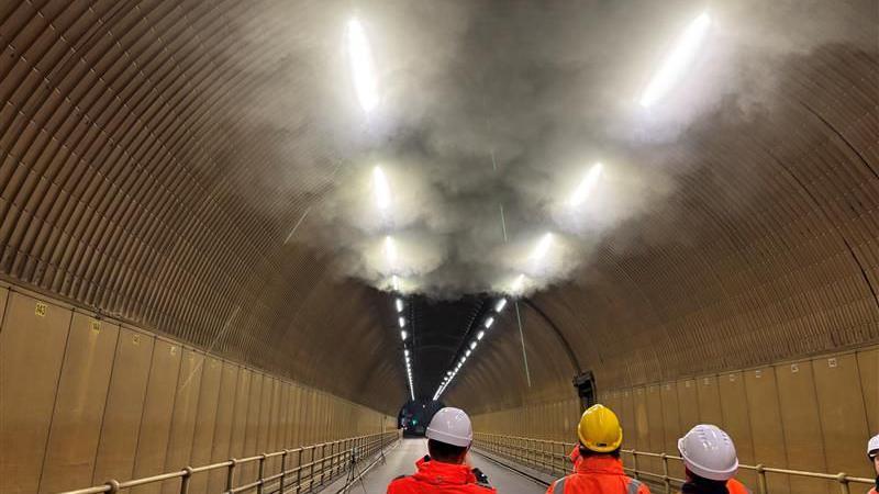 Smoke gathered at the top of a tunnel in Jersey with three people wearing hard hats and orange high vis jackets looking up at it.