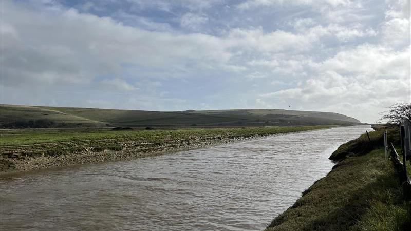 A straight section of the Cuckmere River which leads to the mouth of the river. Banks are on both sides and rolling green hills are in the distance