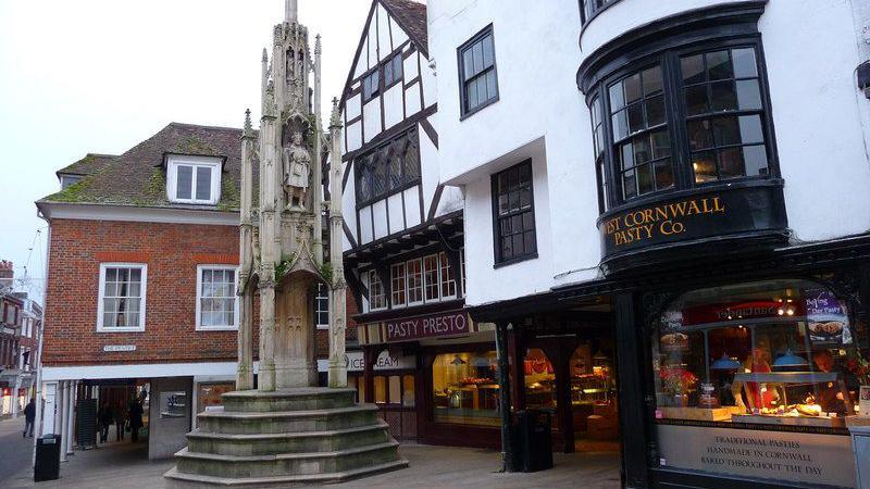 The Butter Cross is a tall, thin, stone structure which is surrounded by shops in Winchester High Street. A pedestal with steps supports a central column and four corner columns. Recessed statues are set halfway up the monument, beneath tapering pinnacles.