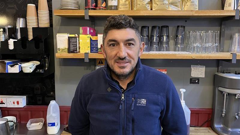 A man behind a cafe counter. He is wearing a blue fleece. He has a salt and pepper beard and hair.