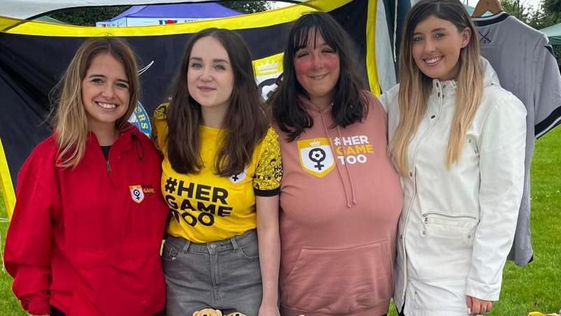 Four women, all with long dark-coloured hair standing under a marquee at a community event. They are all wearing different coloured tops promoting Her Game Too