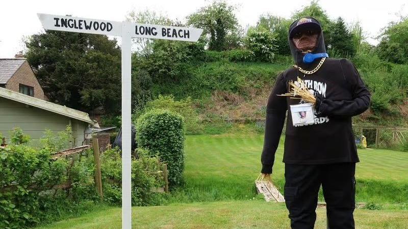 A scarecrow of Dre Dre. It is dressed in a black hoodie and standing next to a sign. It points to Inglewood on the left and Long Beach on the right.