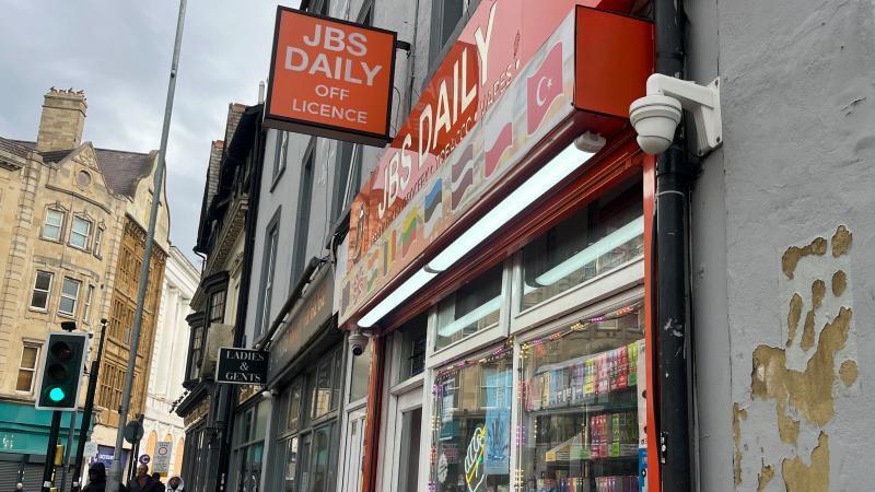 The outside of JB Daily, showing a shop front, traffic lights to the left, people walking along a street, with peeling paint on the wall beside the shop, a downpipe and vapes in the window.