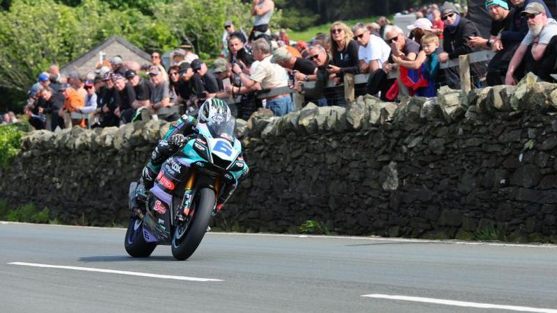 Michael Dunlop, who has the number six on the front of his bike, racing past a stone wall that has several spectators behind in.
