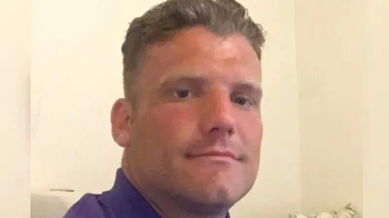Sheldon Jeans has combed back dark hair and a purple polo shirt and is sat in front of a cream wall.