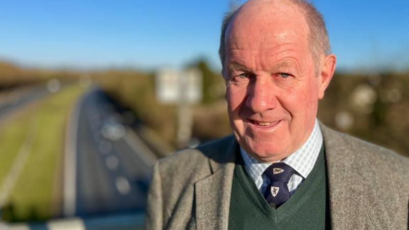 Tim Passmore with very short grey hair, smiling at camera, wearing a grey jacket, green sweater and white shirt with blue tie which has a white loco. He is standing on a road bridge - cars are visible on the road behind him.