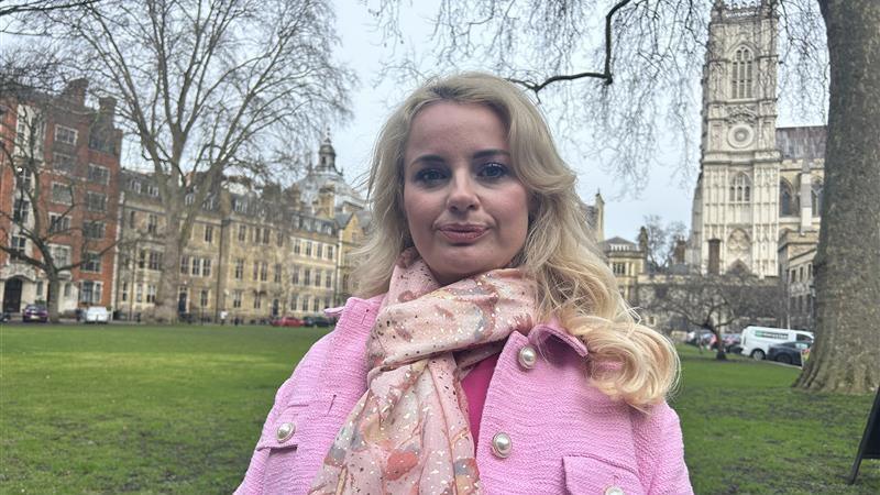 A blonde woman in a pink blouse, scarf and jacket stands on a small park near the Houses of Parliament.