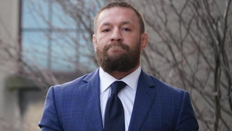 McGregor is wearing a navy suit with a white shirt and dark coloured tie. He has a thick brown beard and short brown hair. He has a straight face looking directly at the camera.