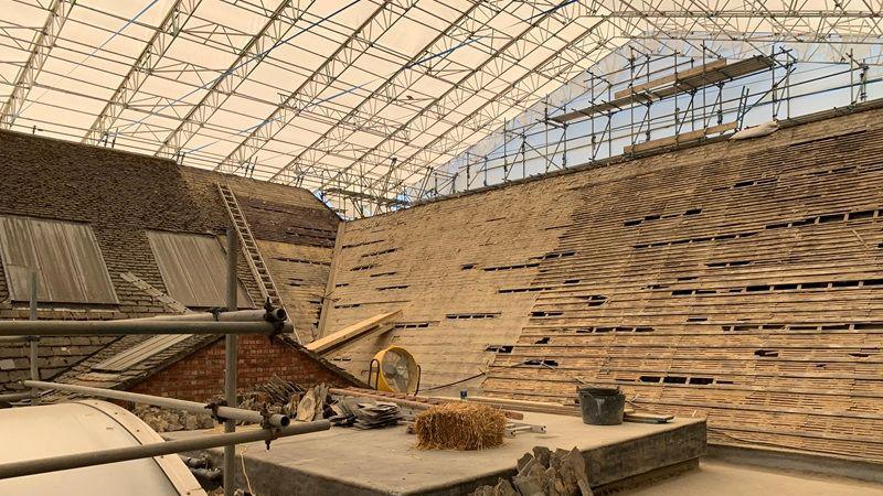 A tent over a slated roof. The roof have several tiles removed. There is also a ladder one of the roof areas.