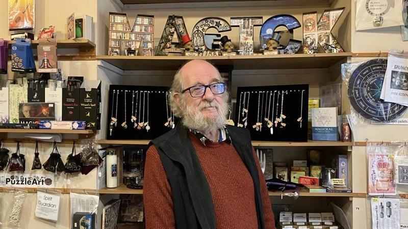 Russell Hall, a man with a white beard and bald head, is wearing a red jumper and black vest. Behind him are wooden shelves filled with various products, including books, tarot cards, candles, and decorative items. At the top shelf, large letters spell out the word “MAGICK”, each letter decorated with different patterns and images.