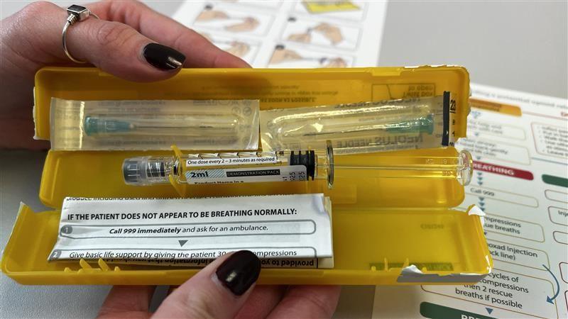 A plastic yellow box is held open showing a syringe with the drug naloxone in it, two needles and an instruction leaflet.