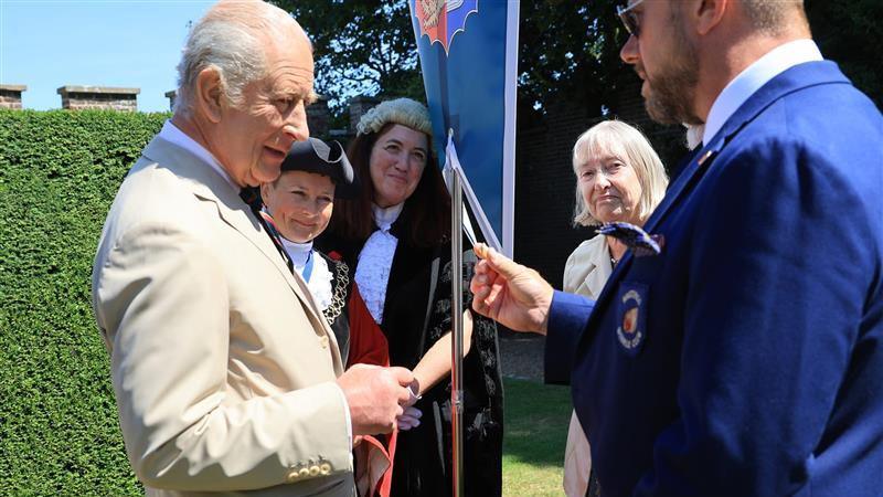 King Charles in a beige suit, stood in a green garden on a sunny day. He is looking at a gold winkle shell being presented to him by a man in a blue suit. Three women, two of which are wearing formal regalia outfits, watch on.