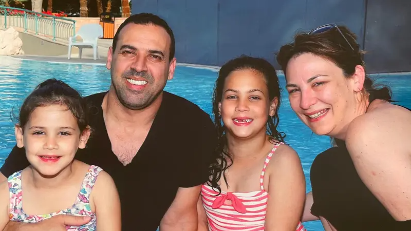 The family of four sitting in front of a swimming pool. Eli holds his younger daughter on his lap, while his other daughter sits next to him, with his wife on the right of the picture leaning in.