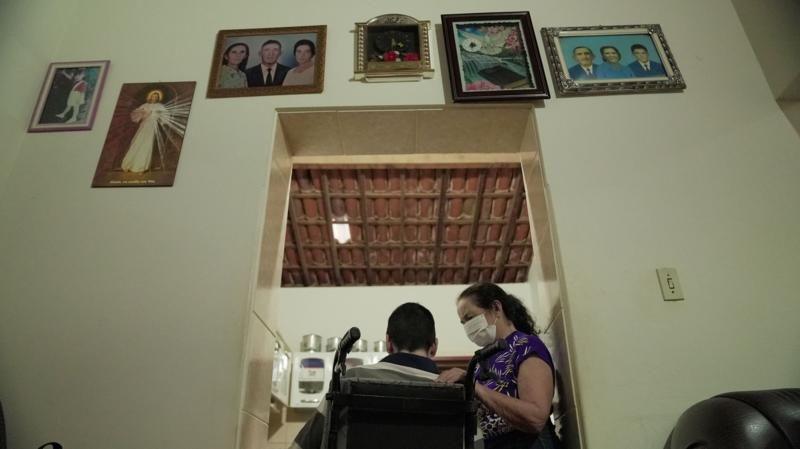 An image showing a man in a wheelchair from behind, being cared for by a woman, with pictures of family members on the wall above him.