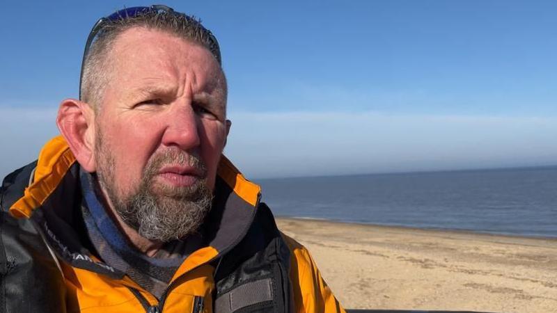 Guy Gibson, the coxswain of Caister Lifeboat, wearing a yellow jacket and lifejacket, with a blue and grey scarf underneath. He has a full beard and moustache and combed short hair. He has sunglasses at the back of his head. He is standing on the deck of the lifeboat, part of which can be seen as a black band on the lower right of the image. The boat is on the beach, with the sand and sea behind, and a clear blue sky.