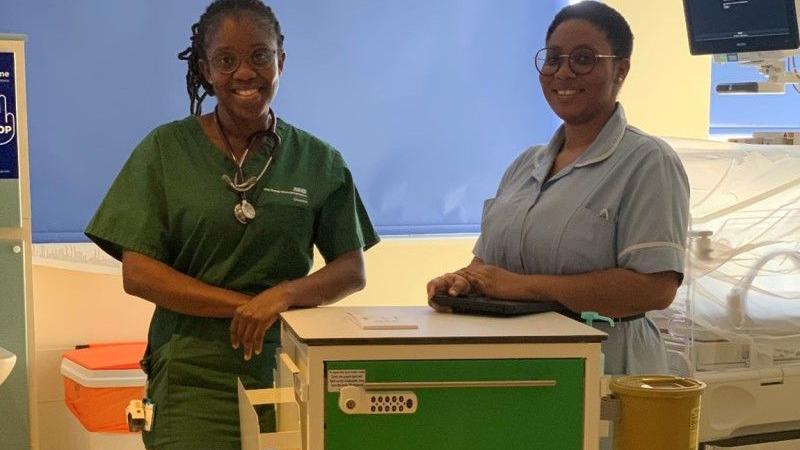 Two women in doctors and nurses scrubs are standing in a hospital ward in front of a bed. They are leaning on a trolley that is cream and has a green drawer. 