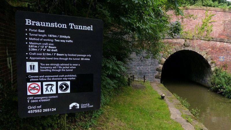A brick tunnel entrance on a canal, with a grass towpath to the left (one mooring post is visible). The tunnel has red brick on the centre and right of the arch, with grey brick on its left. There is a tree to the left of the brick and a black notice outlining details of the tunnel to the left of the tree. The heading on the notice is "Braunston Tunnel".