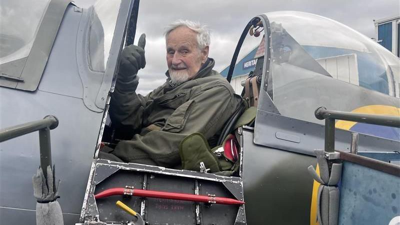 Jack Hemming flying a spitfire in 2024. He is in the spitfire cockpit holding his thumb up in a black glove and wearing an olive green overall.