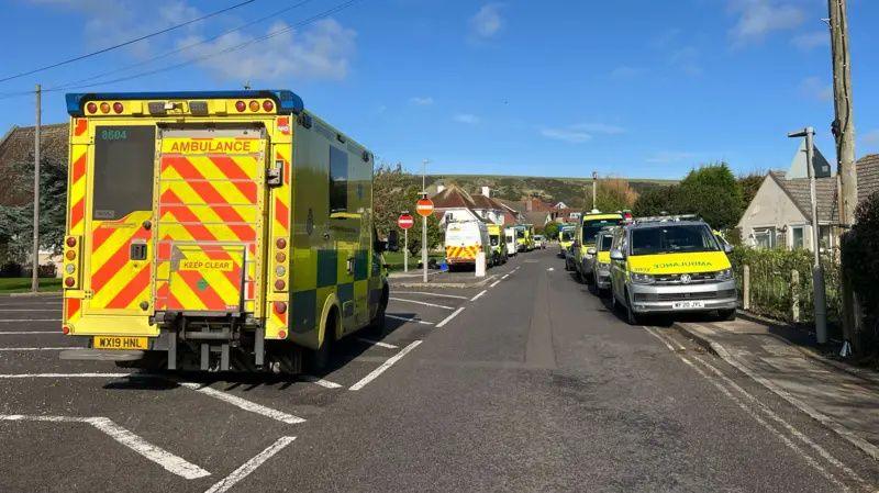 An ambulance parked on a residential road with at least 10 other emergency service vehicles parked on with side of the road stretching back towards the end of the street.