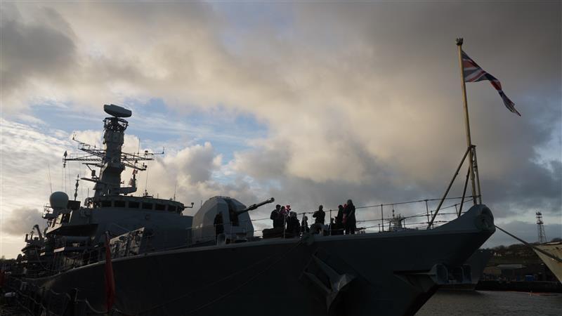 A silhouette of HMS Richmond docking in Plymouth. It is a large frigate and there are silhouettes of people on board. A Union Jack is flying at the front. 