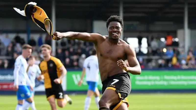 El Bristol Rovers ficha al delantero del Newport, Kamwa. Bobby Kamwa salta con el torso desnudo celebrando tras marcar un gol crucial y sostiene su camiseta con la mano derecha.