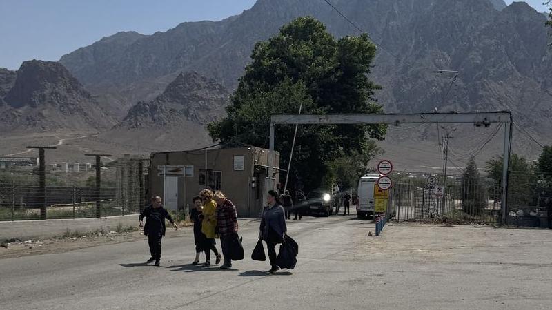 Five people carrying bags walk side by side after passing through the check point at the border crosing with a view of mountains behind them