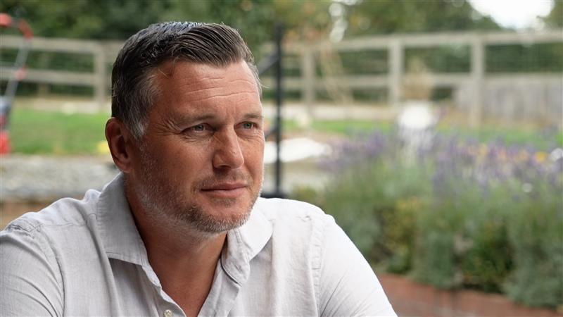 Andy Marshall, in a grey T-shirt, looking towards the camera with a garden in the background. He has brown hair with stubble. His head and shoulders are visible in the shot.