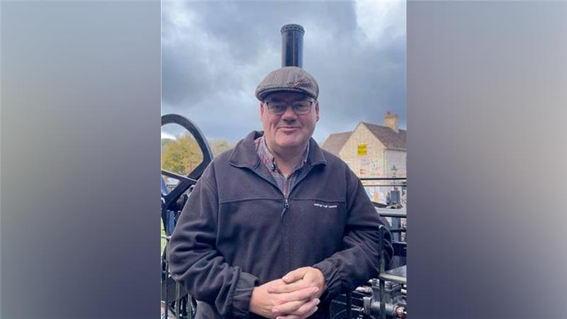 A man wearing a brown flat cap, glasses and a black zip fleece is standing in front of a small black steam engine with a tall chimney.
