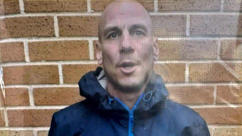 A bald man, who is wearing a dark blue coat with the hood down, has his mouth partially open as he stands in front of a wall.