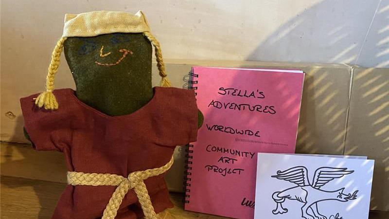 A 30cm (1ft) tall hand puppet made of linen with a long burgundy dress. a yellow hat and pigtails sticking out at each side, it also has a yellow belt tied in the middle. Beside the puppet is a pink book saying community art project and a drawing of a dragon like creature in black felt tip on a white card background.
Behind is a brown cardboard parcel, that all the items were sent in.