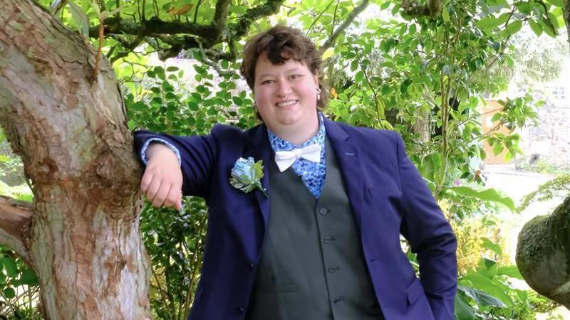 Freddy Morris in a three-piece suit with a purple jacket and a white bow tie, smiling and leaning against a tree. She has neck-length brown hair. 