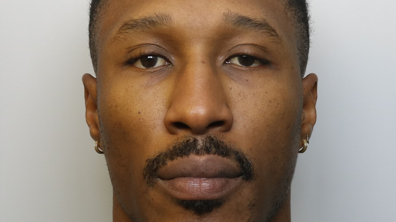 A man looking at the camera in a police mugshot, with a moustache and wearing two gold earrings.