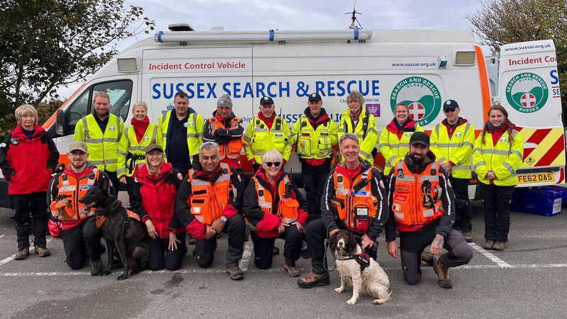 Brown and white springer spaniel Millie with her handler Darren Yeates and a group of 16 other members from the organisation Sussex Search and Rescue. They are lined up in two rows, in high viz yellow and orange coats, in front of a white van with the name of the organisation along its side.