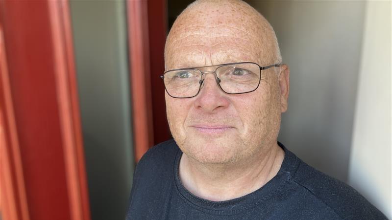 A bald man wearing glasses and a black t-shirt. He is stood in front of a red door.