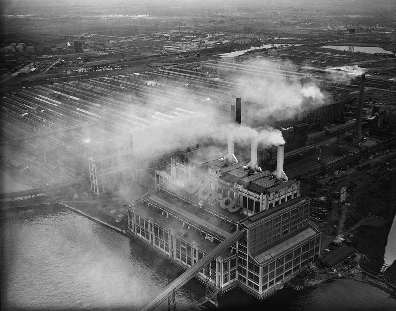 A black and white photograph of Ford Motors car factory on the River Thames at Dagenham, London, on December 7th 1960. Smoke is being pumped out of the factory's chimneys and covers the sky