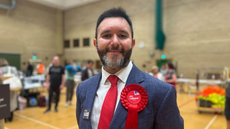 Image of Sadik Al-Hassan. He has dark hair and facial hair, and is wearing a blue suit, white shirt, red tie and a red labour badge.